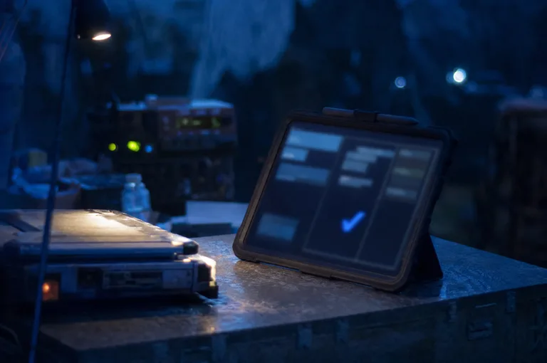Rugged tablet propped on a field table at night, its screen showing blurred interface rectangles and one glowing blue check shape