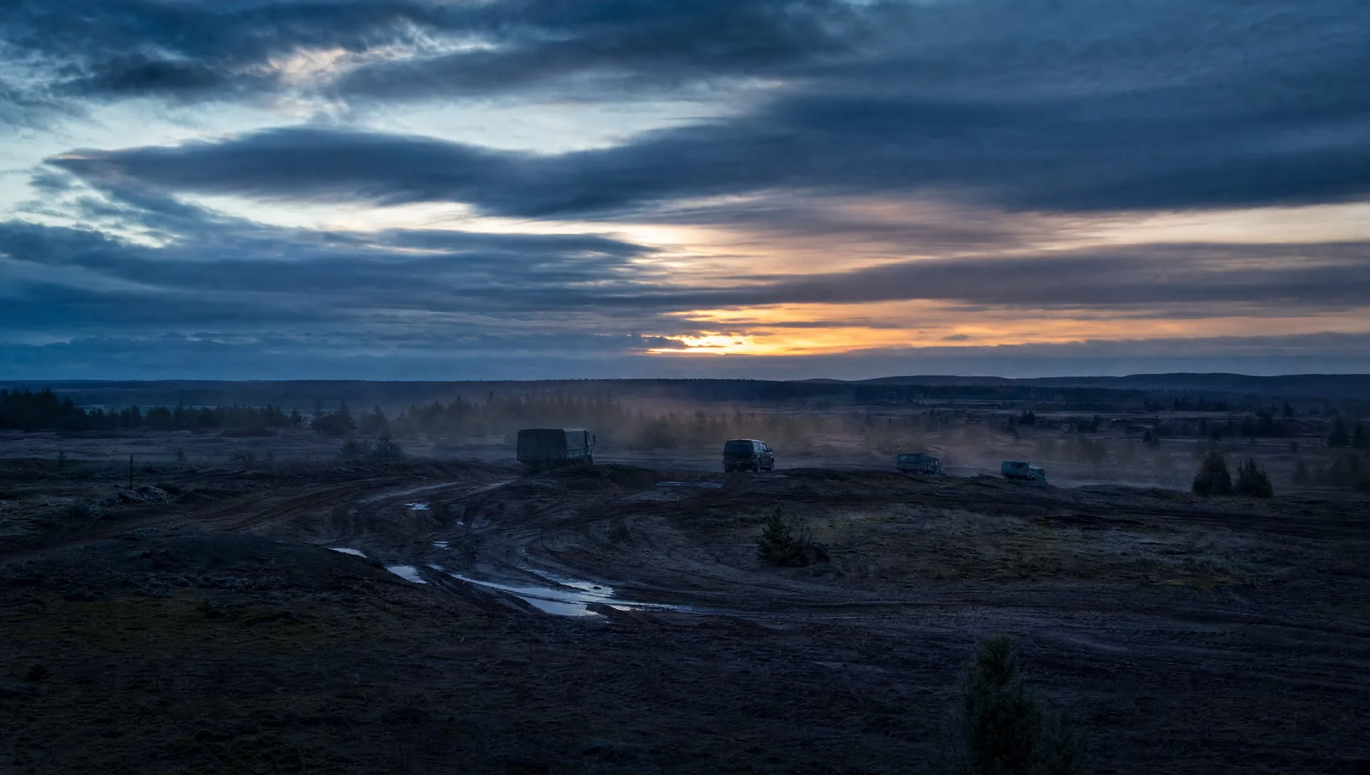 Golden hour over unpopulated training terrain with tire tracks in mud, unmarked vehicles in the middle distance under a dramatic sky
