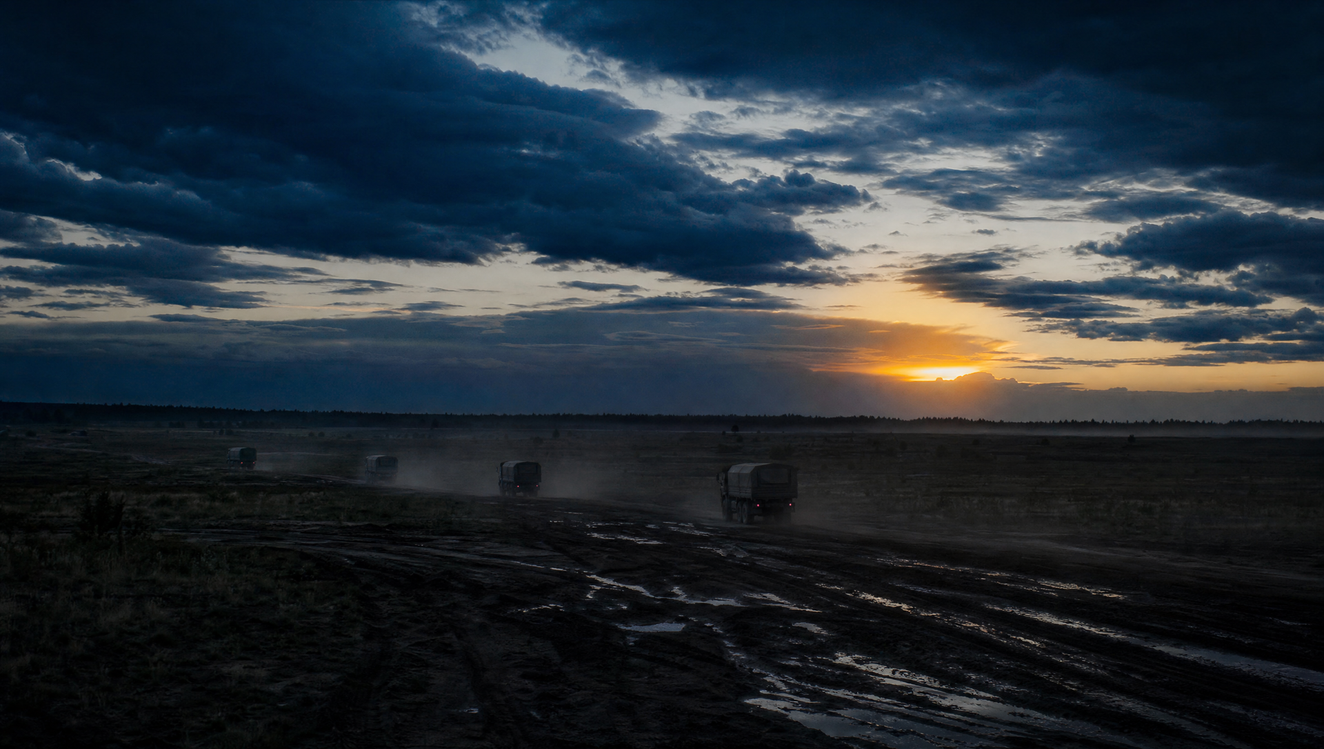 Open training range at golden hour with rutted mud tracks catching low sun, unmarked vehicles far in the distance under a vast dramatic sky